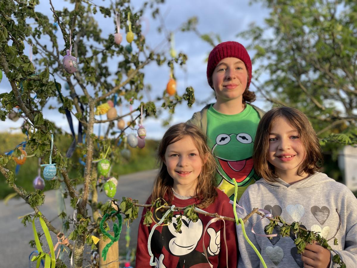 Three smiling girls stand beside a tree decorated with painted eggs